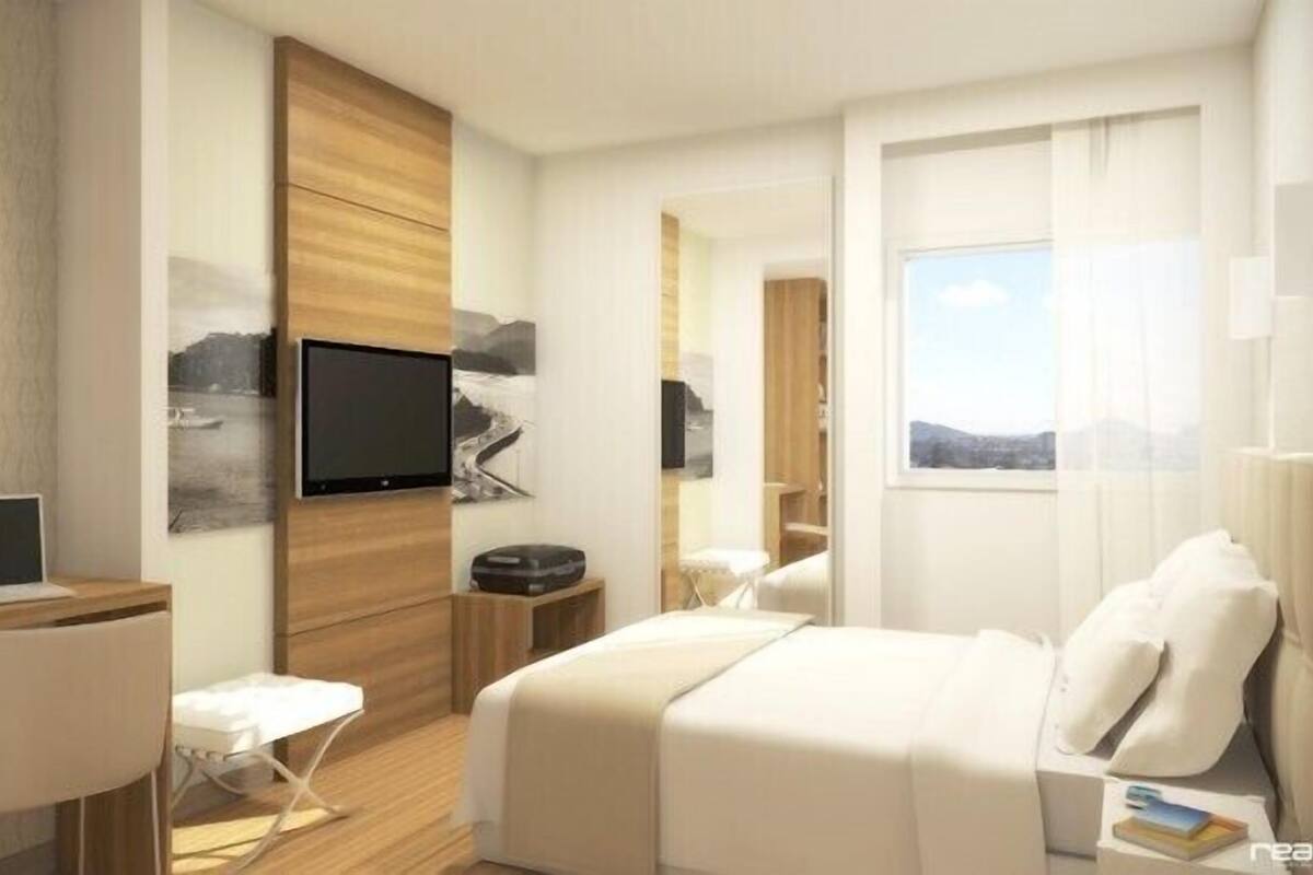 A modern hotel room with a bed, a desk with a laptop, a TV mounted on the wall, and a window with a view.