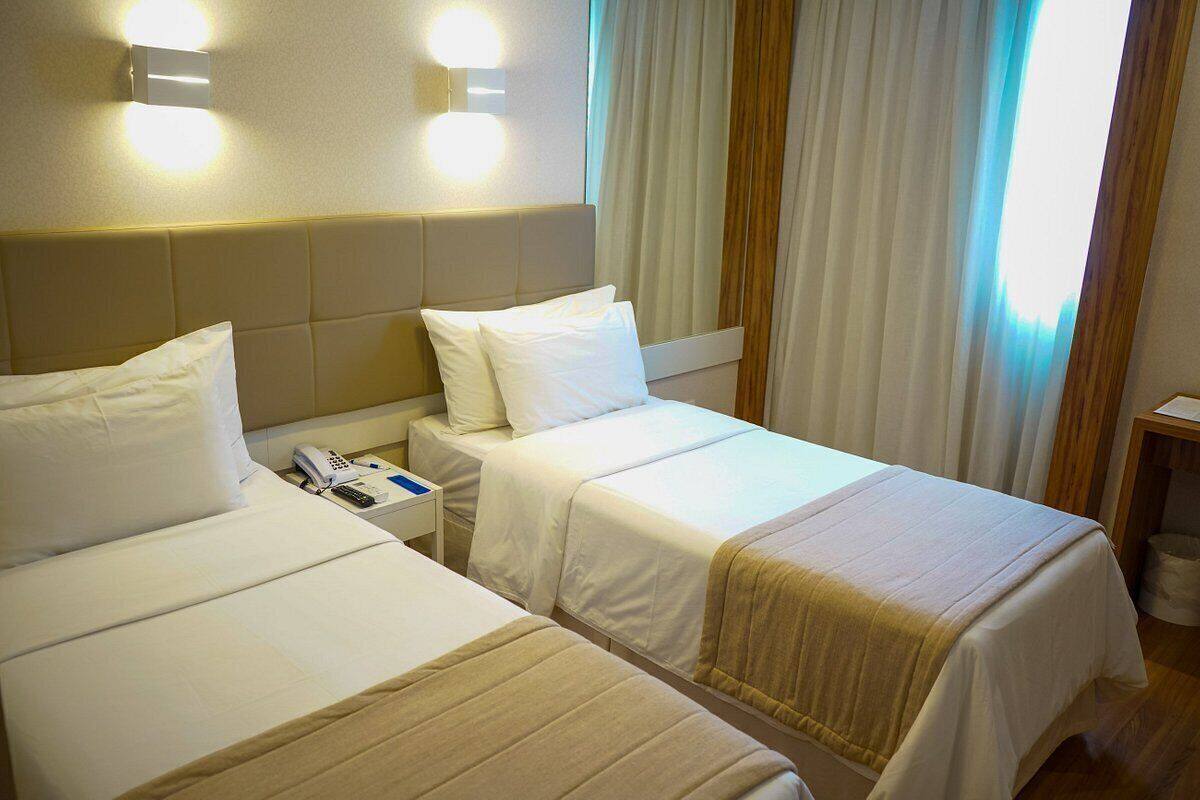 A hotel room with two beds, a bedside table with a phone, and a wall-mounted light fixture.