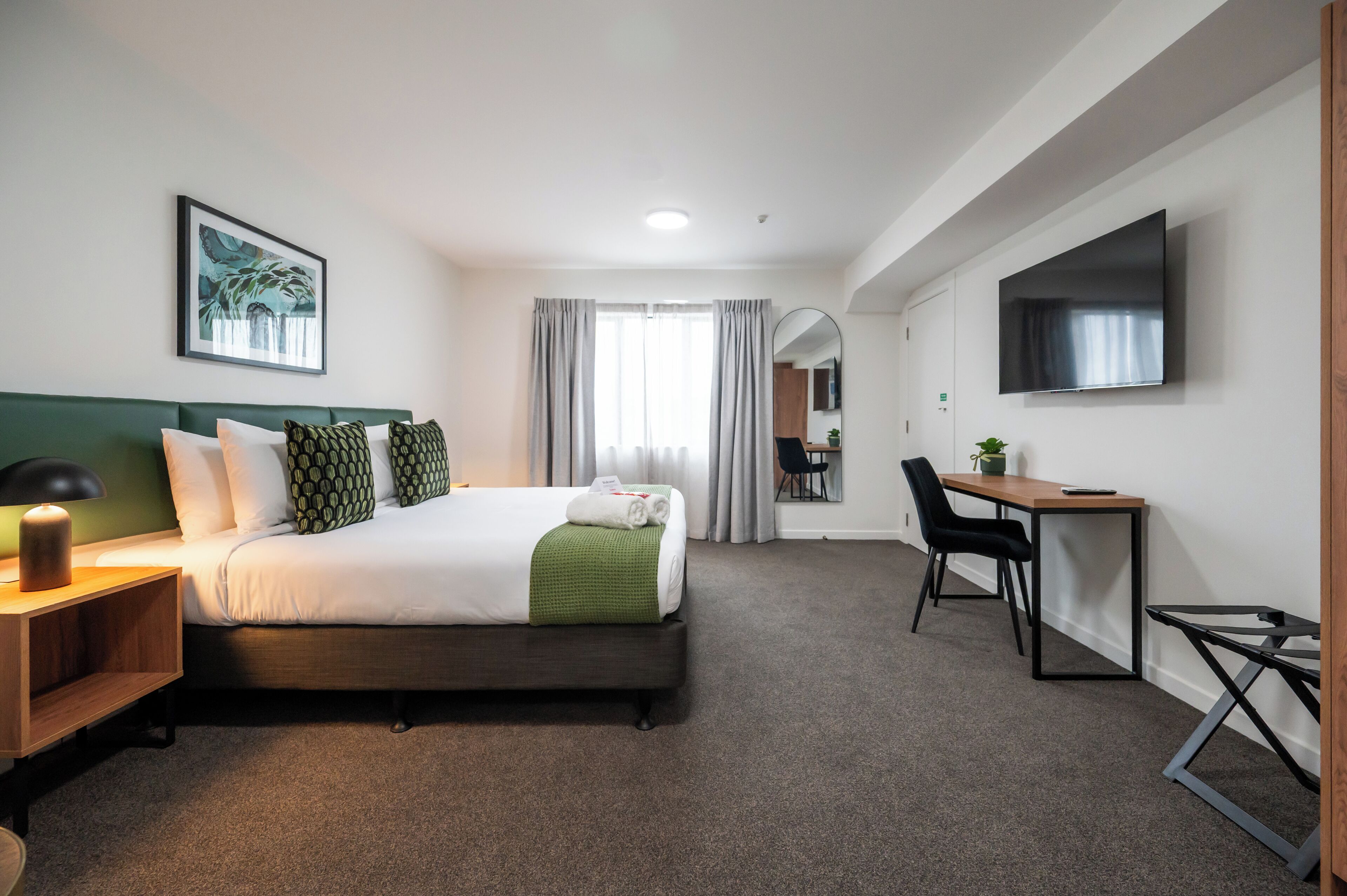 A hotel room with a bed, a desk, a chair, a TV, and a painting on the wall.