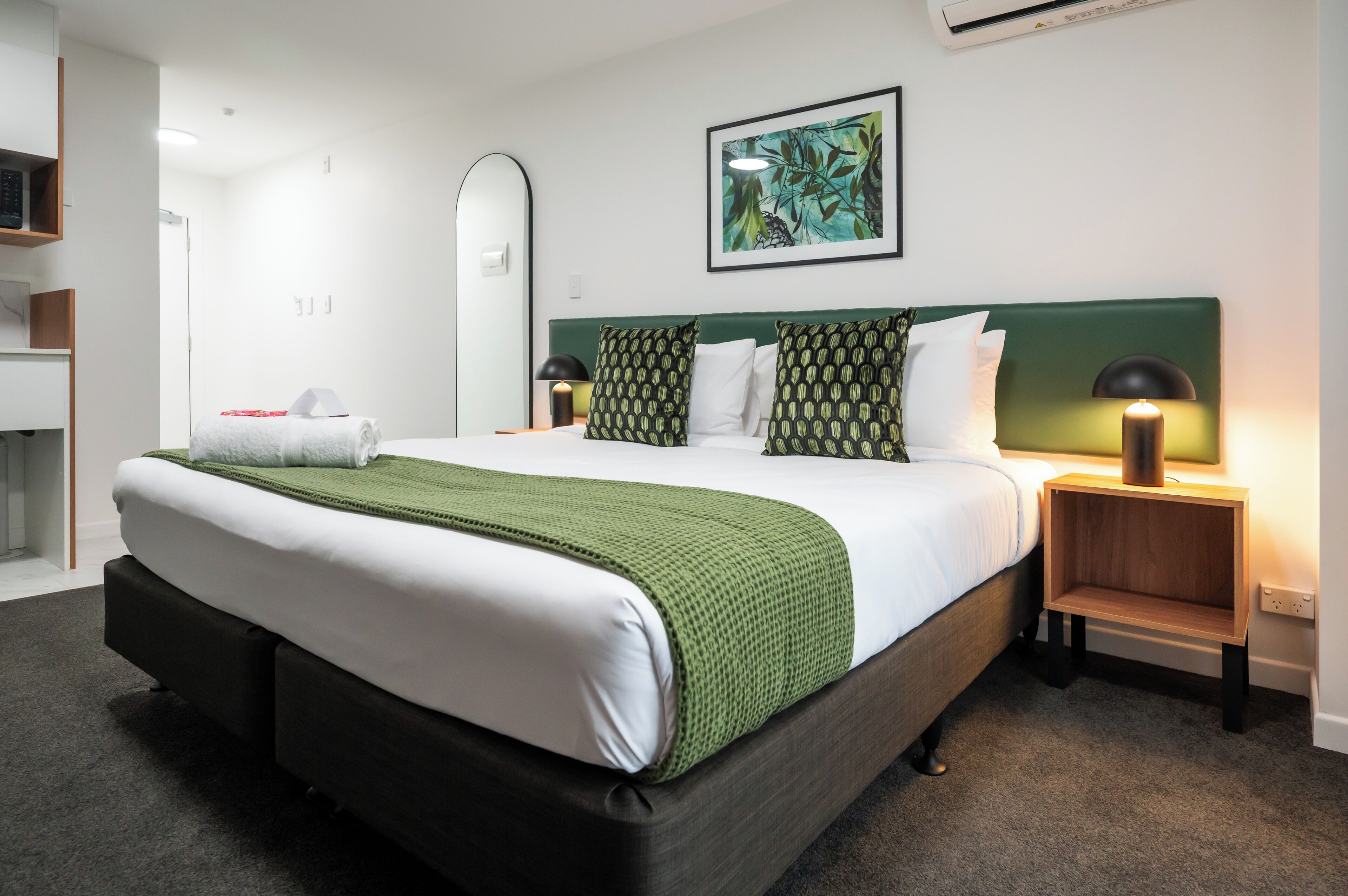 A neatly made bed with a green throw, two bedside tables with lamps, a mirror, and a framed artwork on the wall.