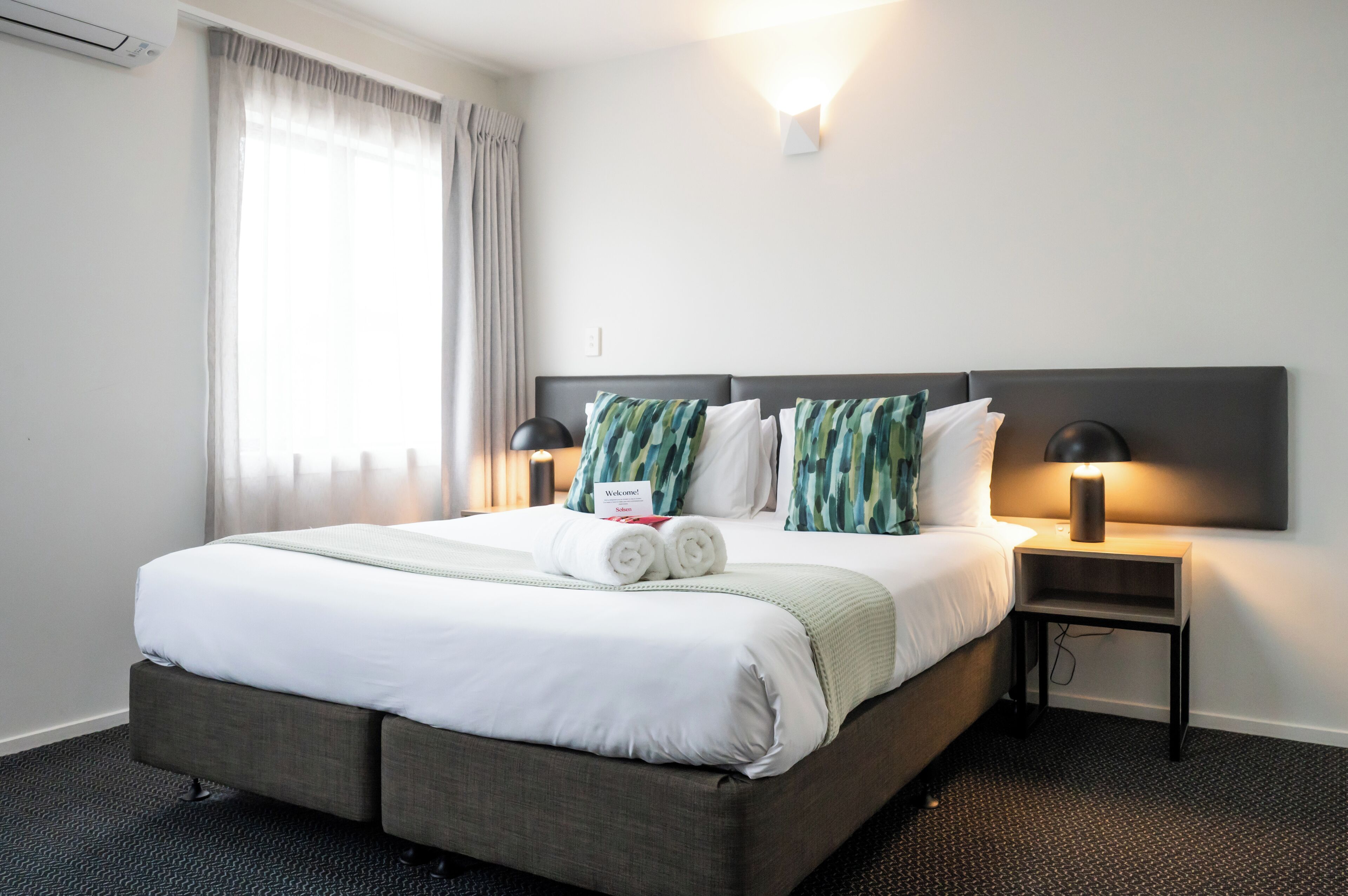 Una cama bien hecha con sábanas blancas y almohadas verdes y blancas, una mesita de noche con lámpara, y una ventana con cortinas.