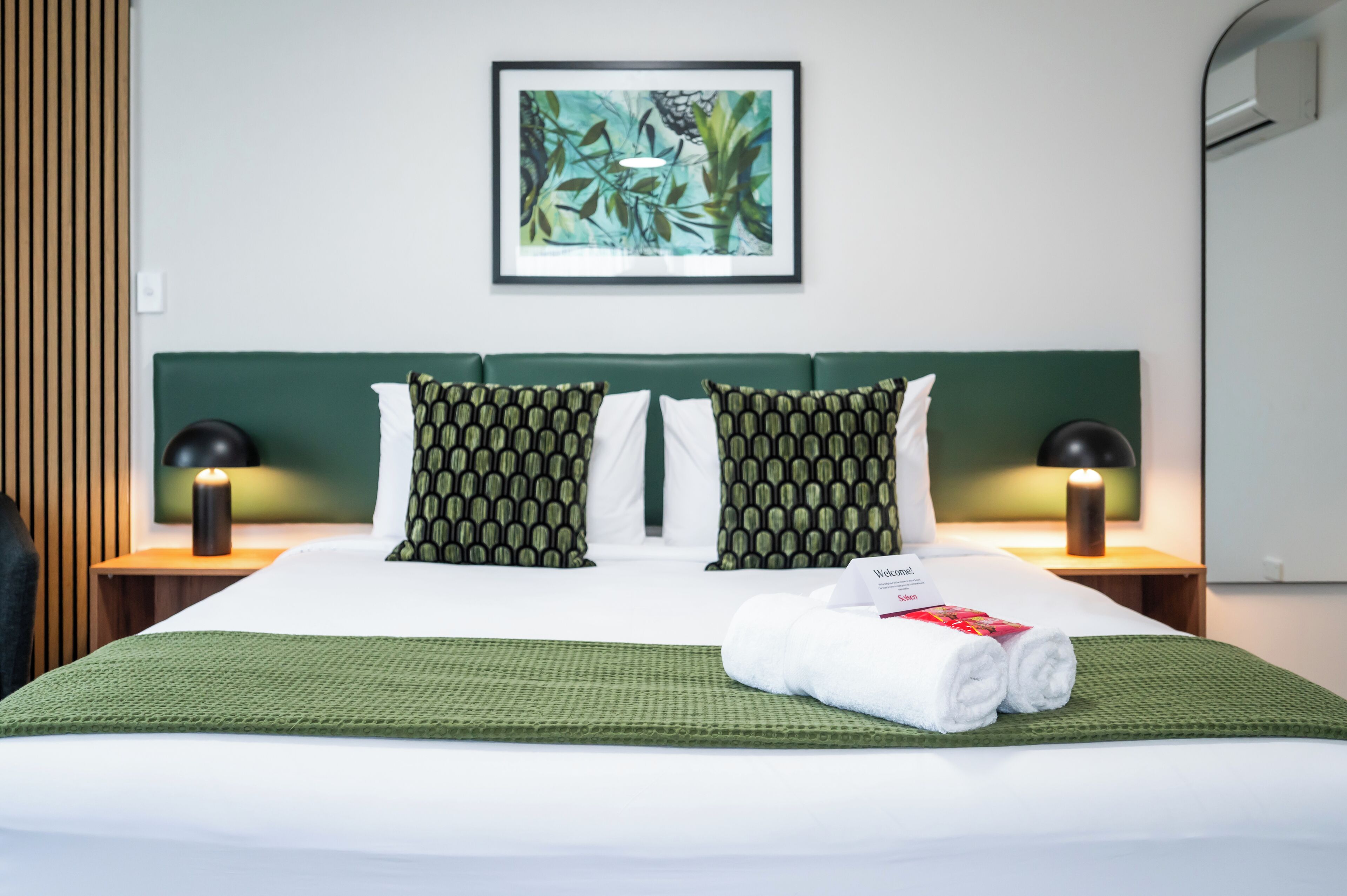 Una cama bien hecha con ropa de cama verde, dos almohadas estampadas, una obra enmarcada encima y una mesita de noche con una lámpara.