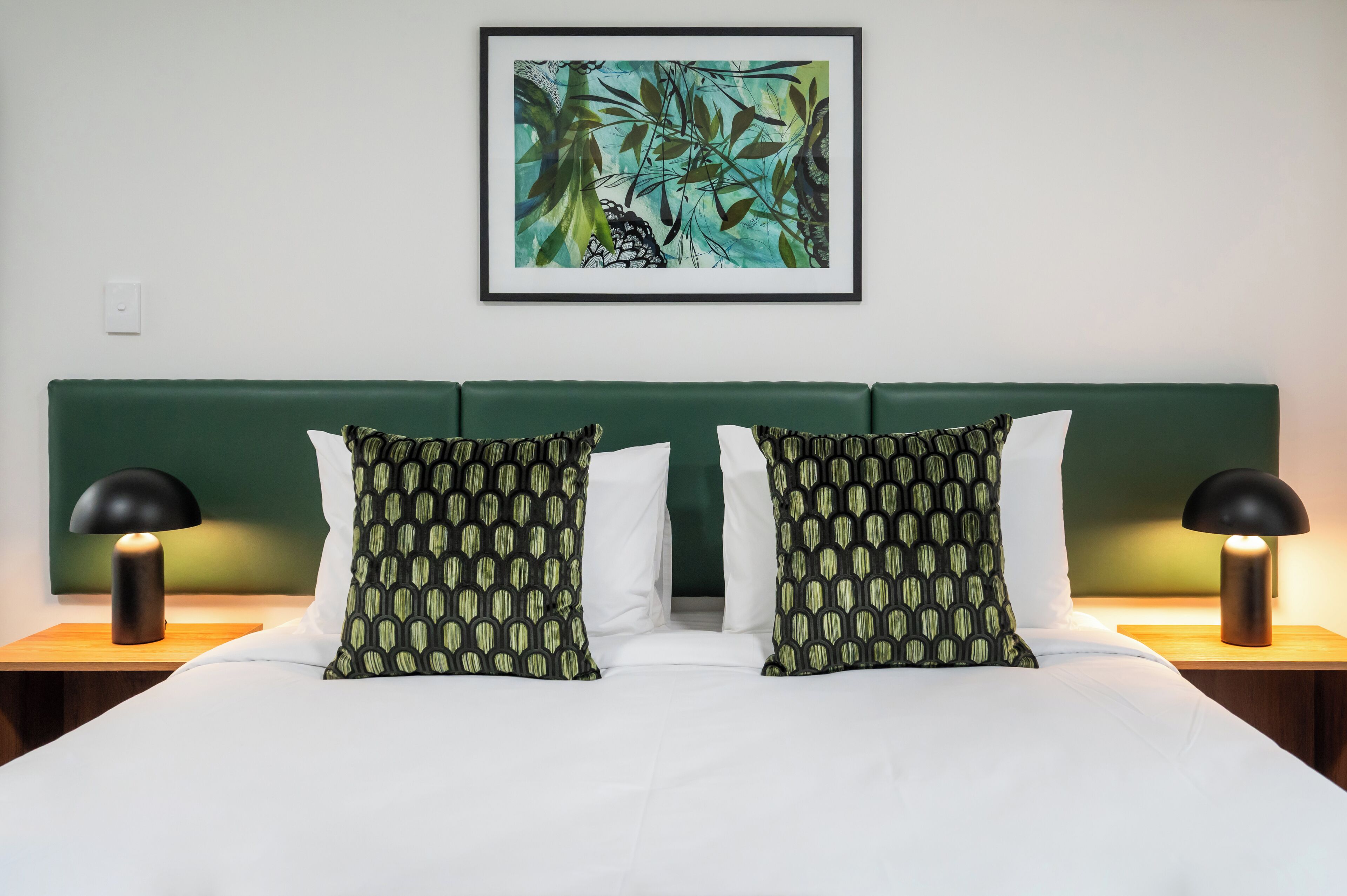 A double bed with green and white bedding, two patterned pillows, a green headboard, and a framed abstract painting above.