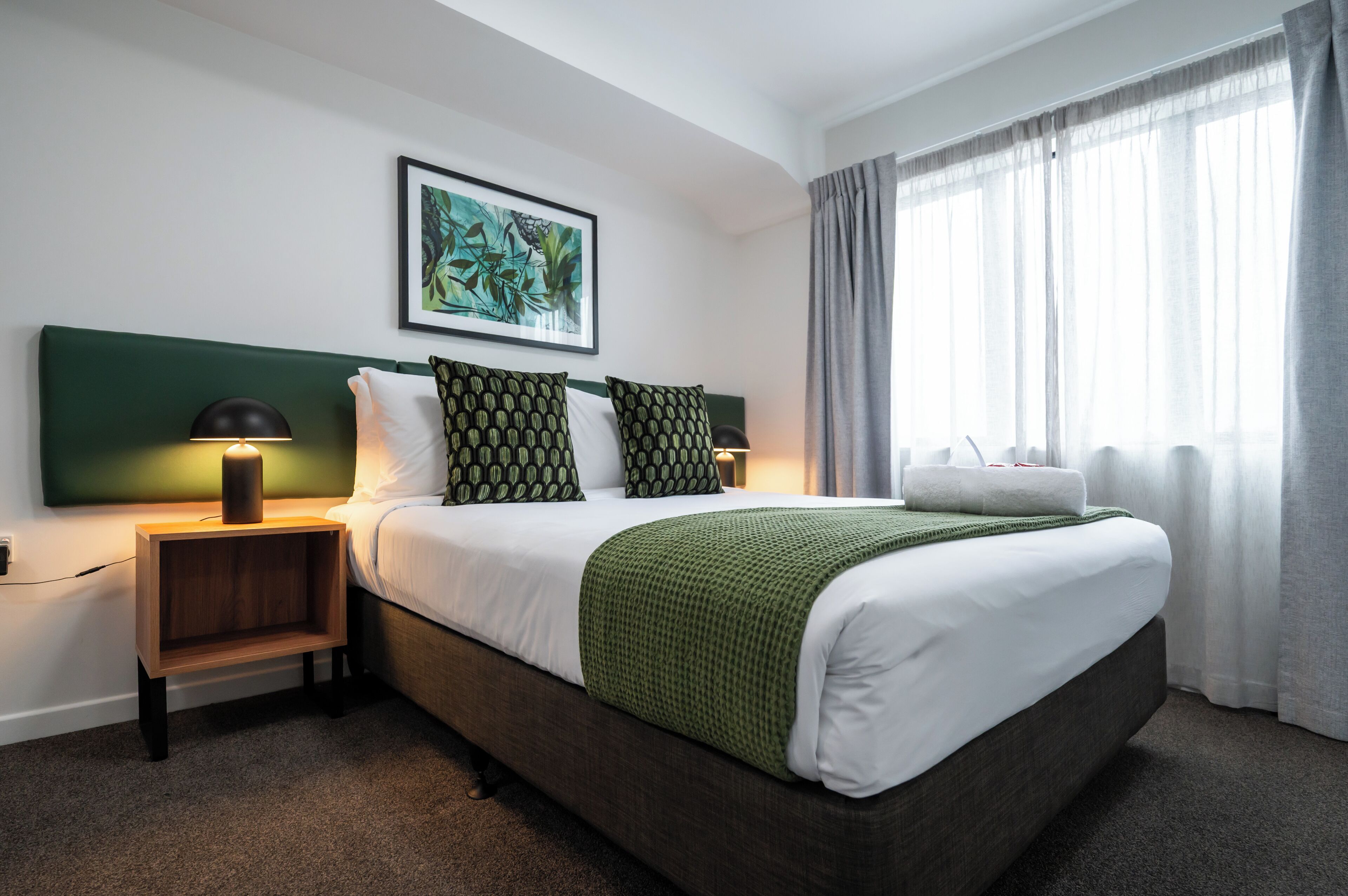 A neatly made bed with a green throw, a nightstand with a lamp, and a framed artwork on the wall.