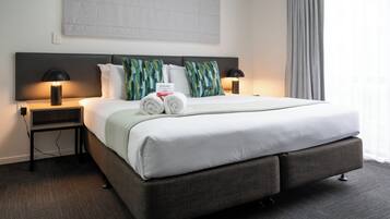 A neatly made bed with white linens, two pillows, and a welcome sign. There are two bedside tables with lamps, a grey headboard, and a grey curtain on the window.
