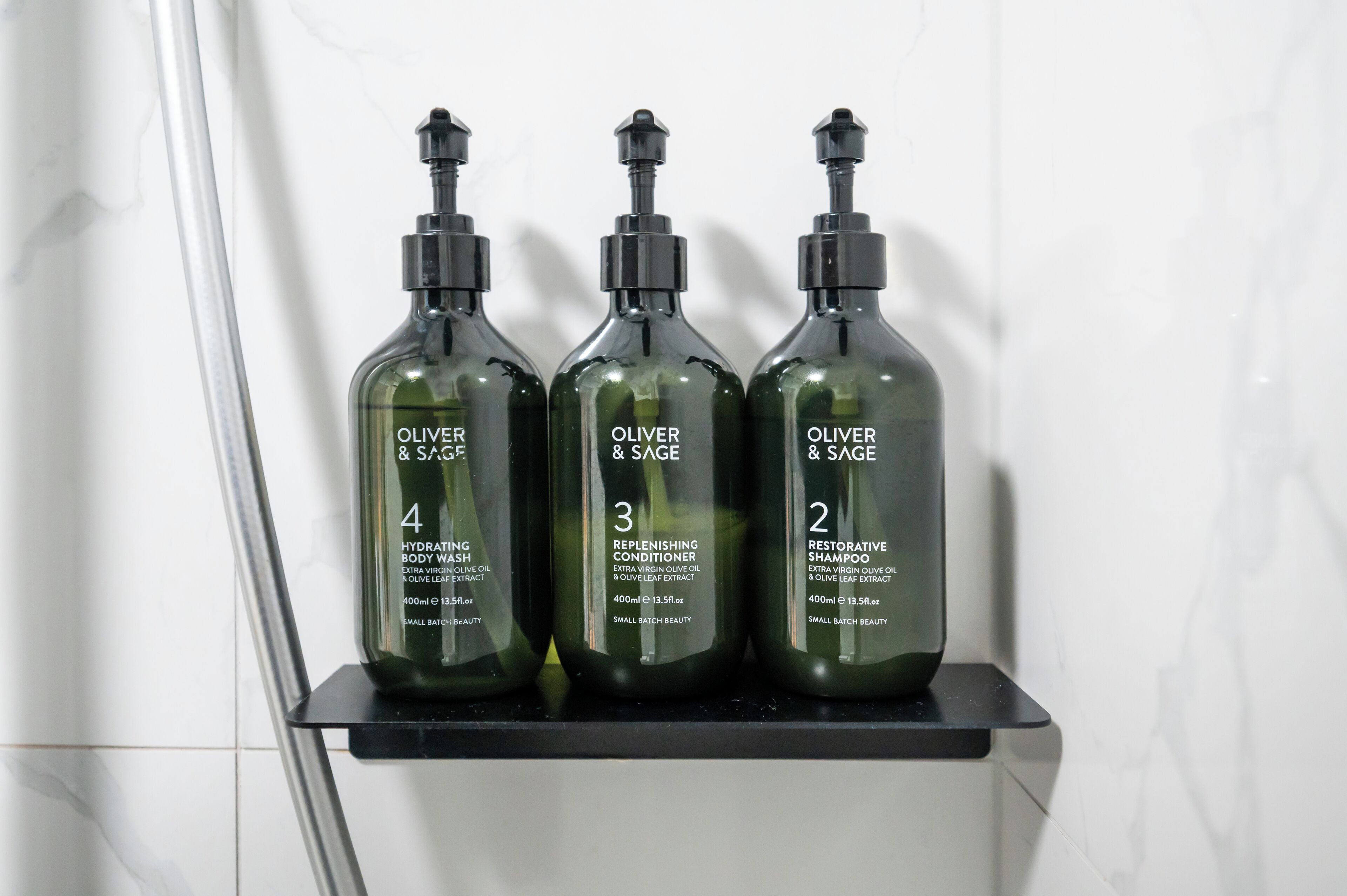 Three bottles of Oliver & Sage body wash on a black shelf against a white wall.
