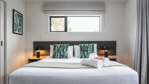 Un cuarto de hotel con una cama grande, dos mesitas de noche con lámparas, un ventanal con vistas y un cuadro en la pared.