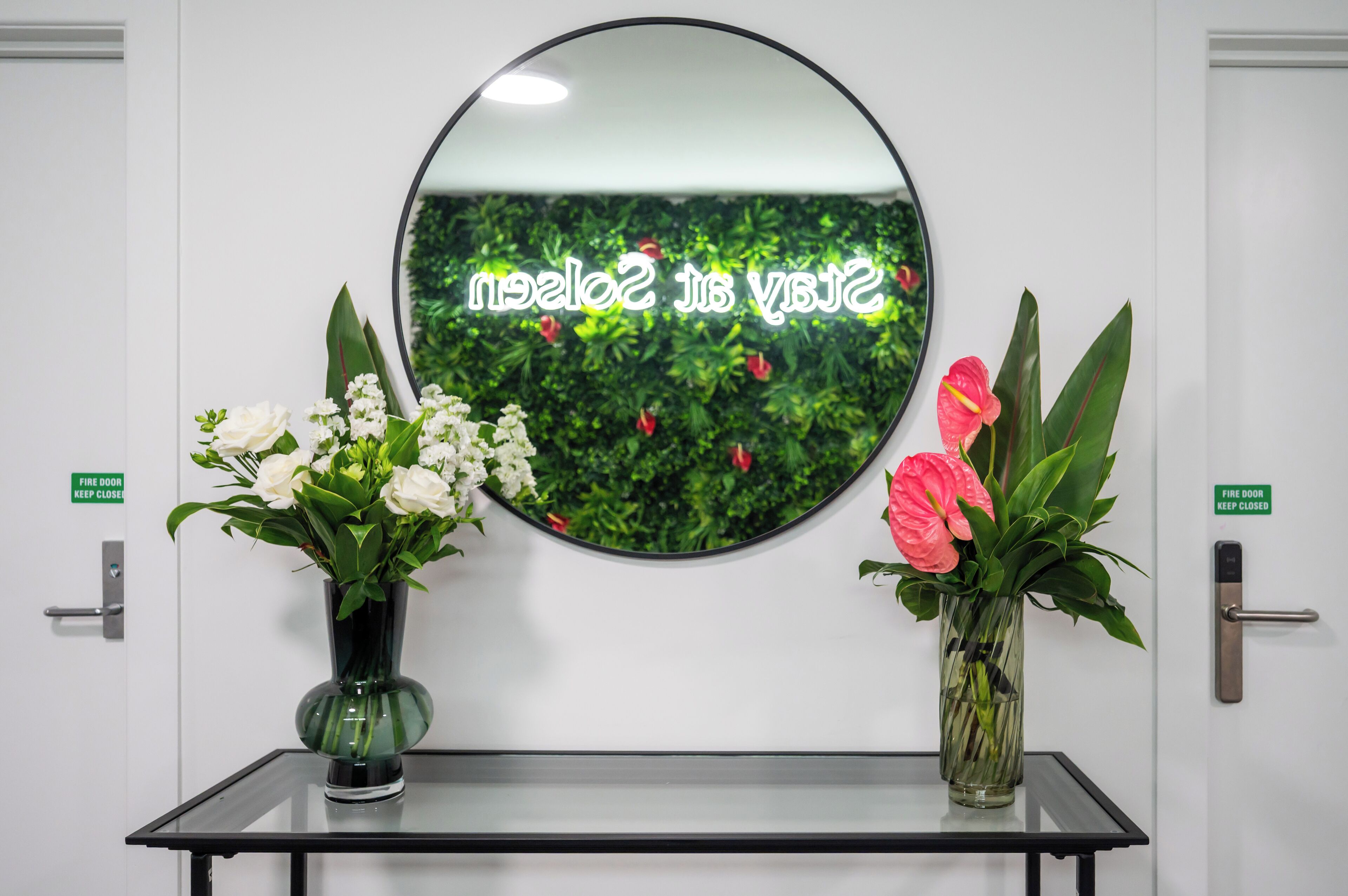 Een receptiegedeelte met een ronde spiegel, twee vazen met bloemen en een bordje waarop staat dat de branddeur gesloten moet blijven.