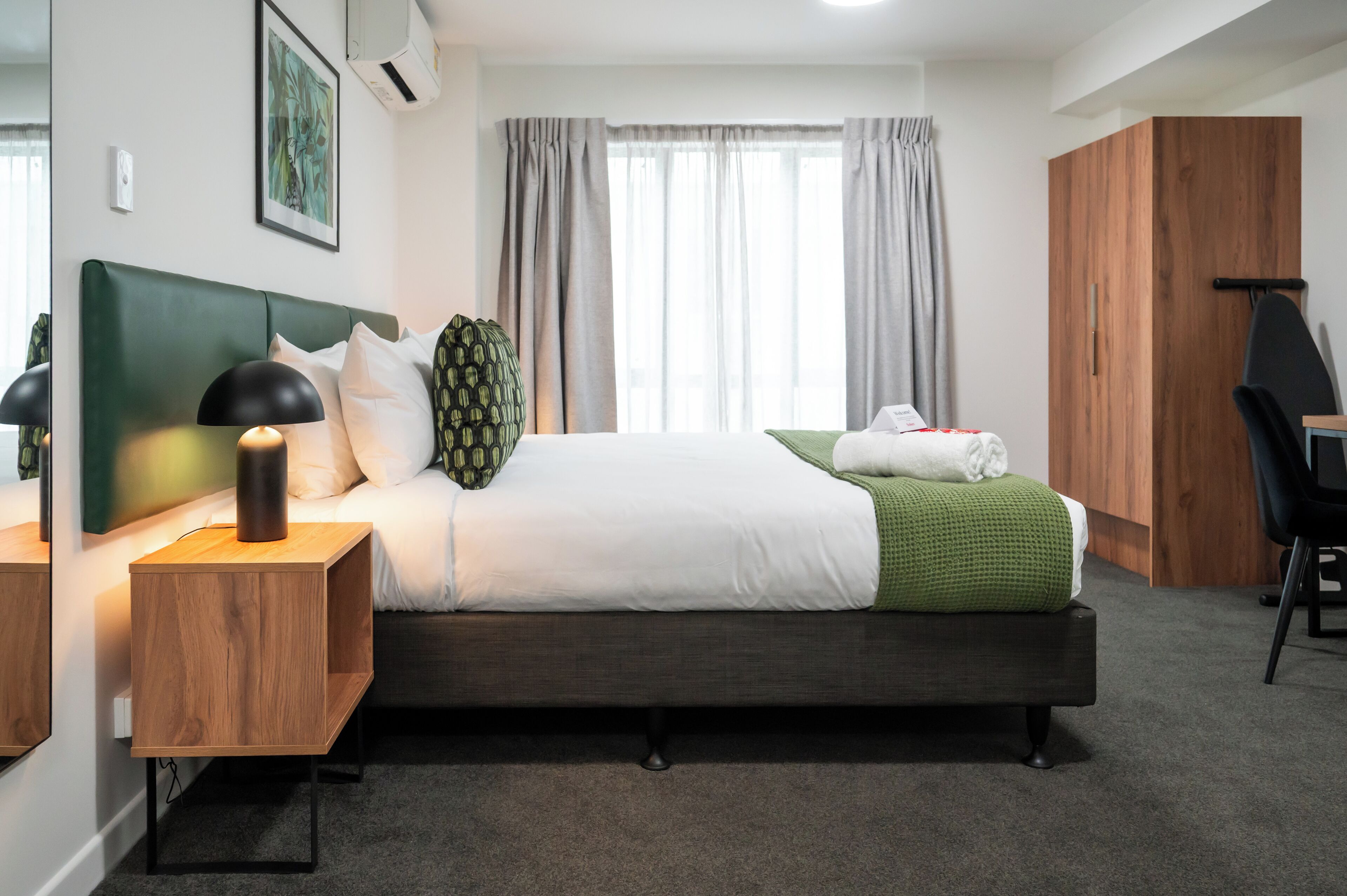 A modern hotel room with a large bed, a wooden nightstand, a desk, and a wardrobe.