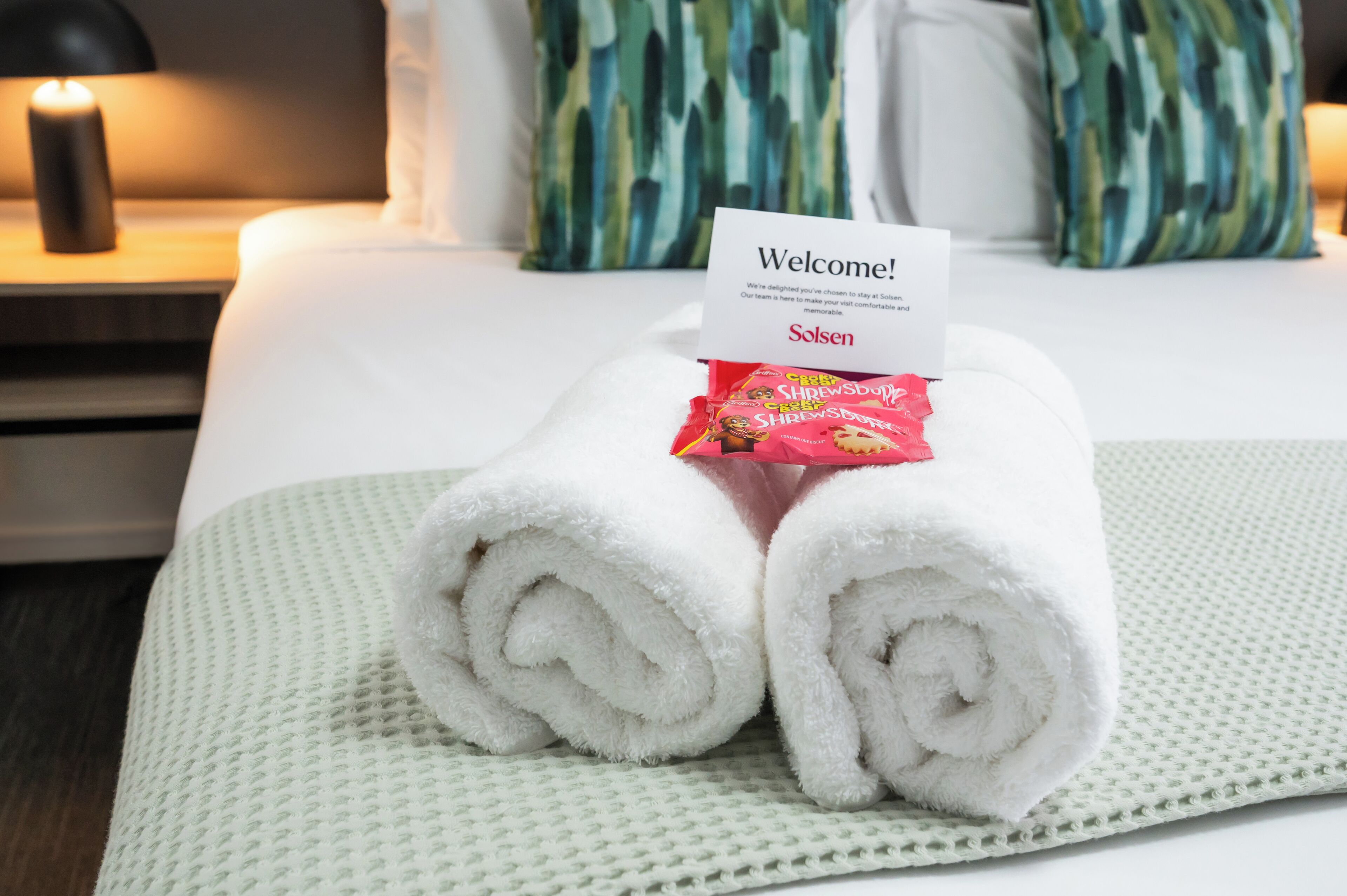 A neatly made bed with a welcome sign, a rolled white towel, and a packet of Shrewsbury Shredded Chicken.