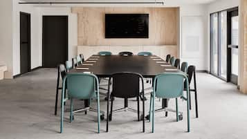 Un salón de conferencias con una mesa larga, sillas color turquesa, una televisión montada y ventanas grandes.