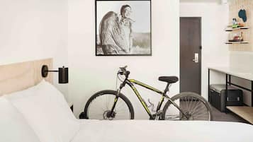 Una habitación de hotel con una cama, una bicicleta, un cuadro en la pared y un armario con estantes.