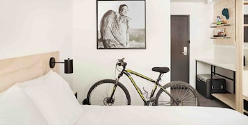 Una habitación de hotel con una cama, una bicicleta, un cuadro en la pared y un armario con estantes.
