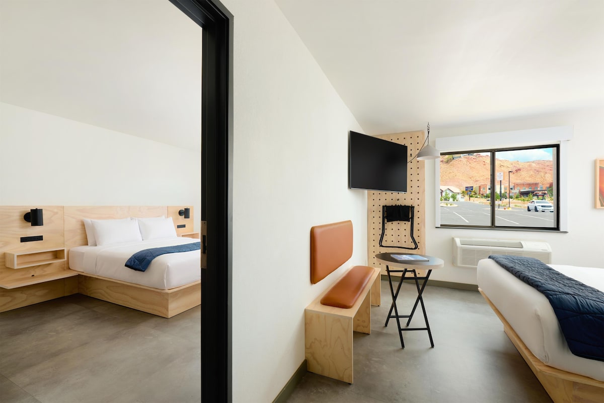 A modern hotel room with a bed, a desk, a chair, a TV, and a window with a view of a parking lot and buildings.