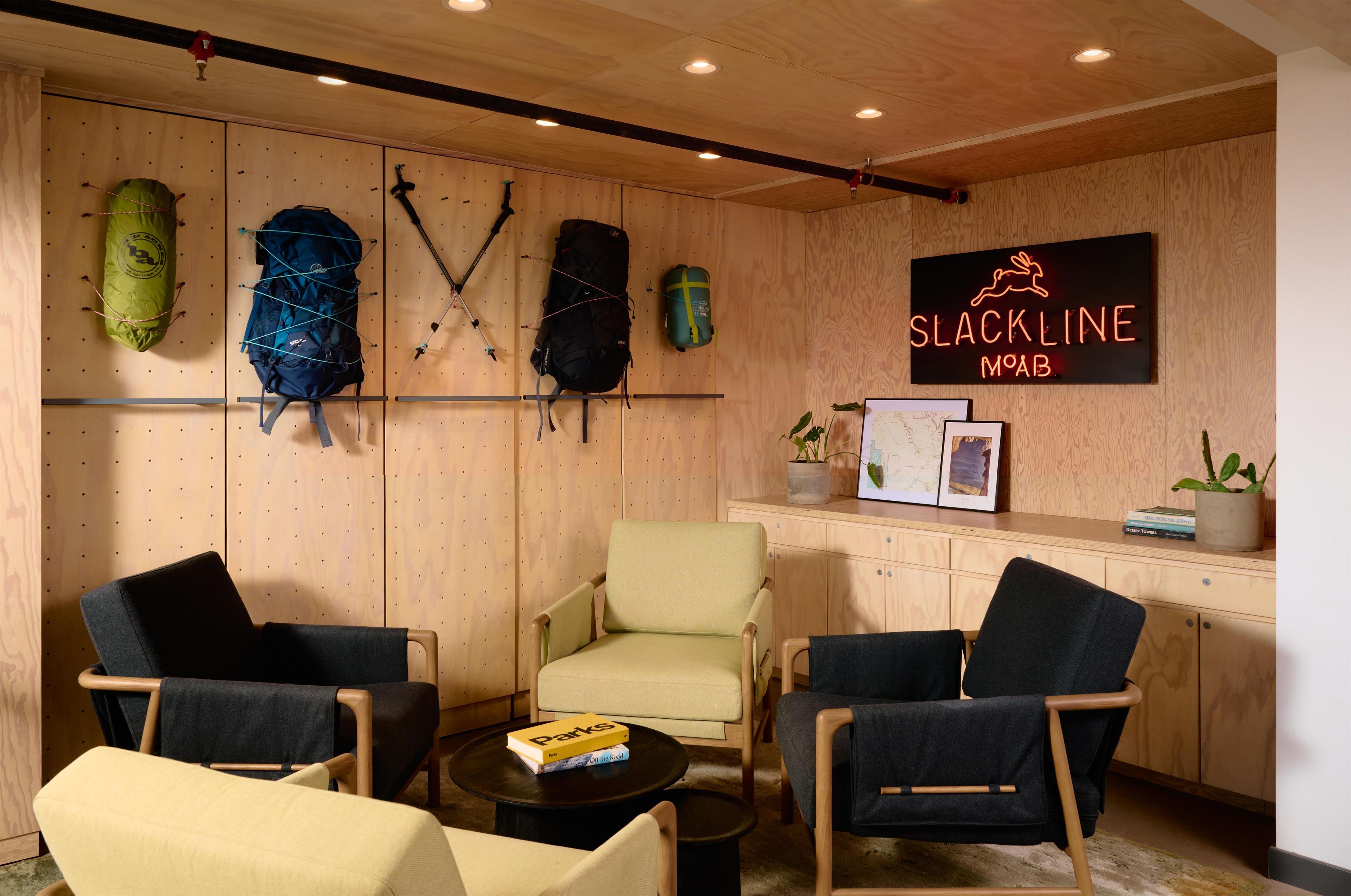 Una habitación con paneles de madera, un letrero de neón que dice 'SLACKLINE MOAB' y varias sillas y mochilas.