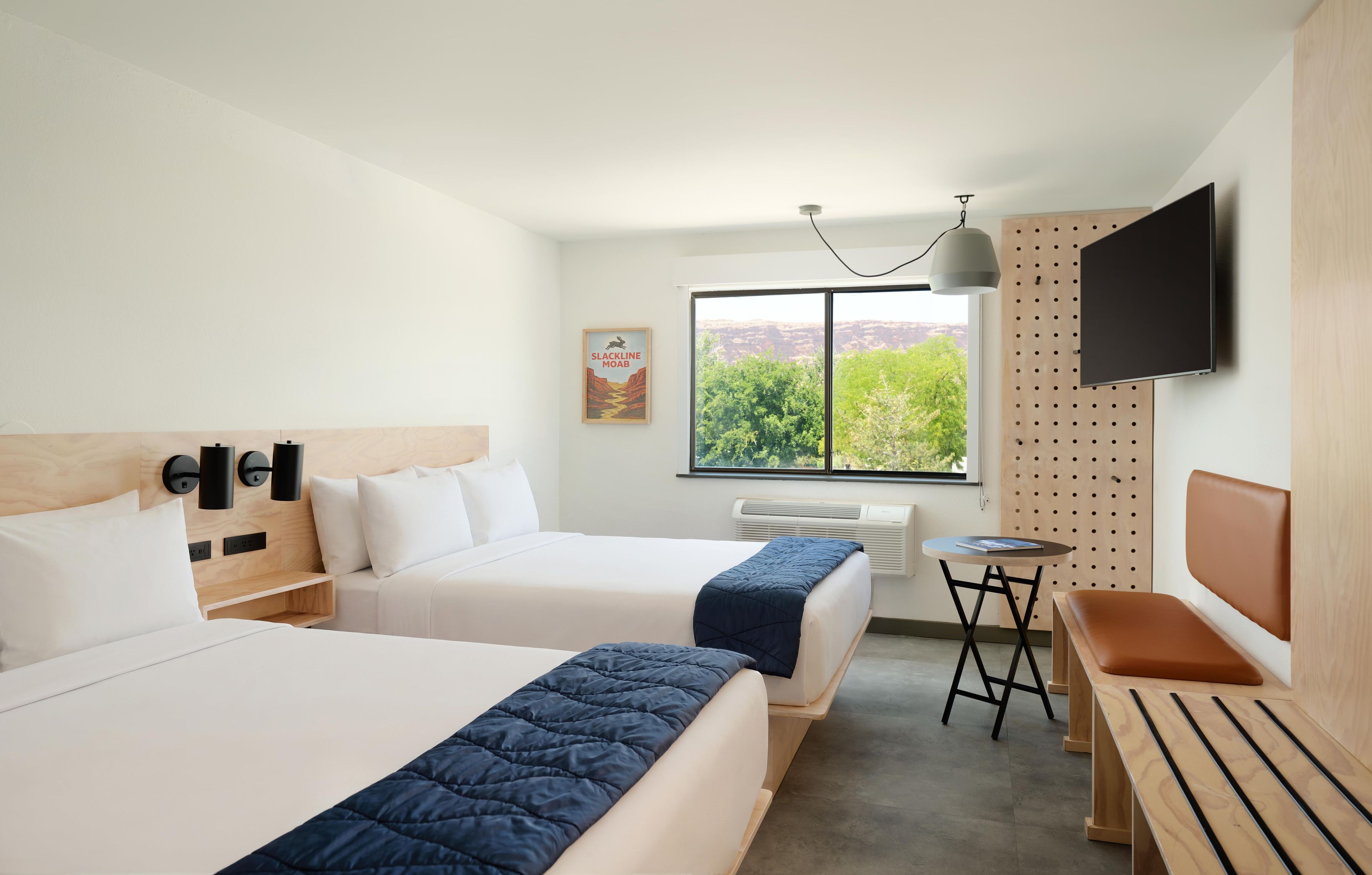 A hotel room with two beds, a desk, a chair, a TV, and a window with a view of greenery.