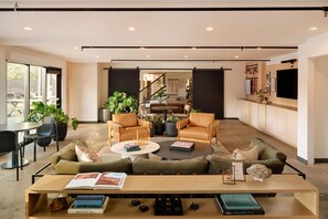 A spacious living area with a sofa, coffee table, and chairs, a dining table with books, and a kitchen area with a TV.