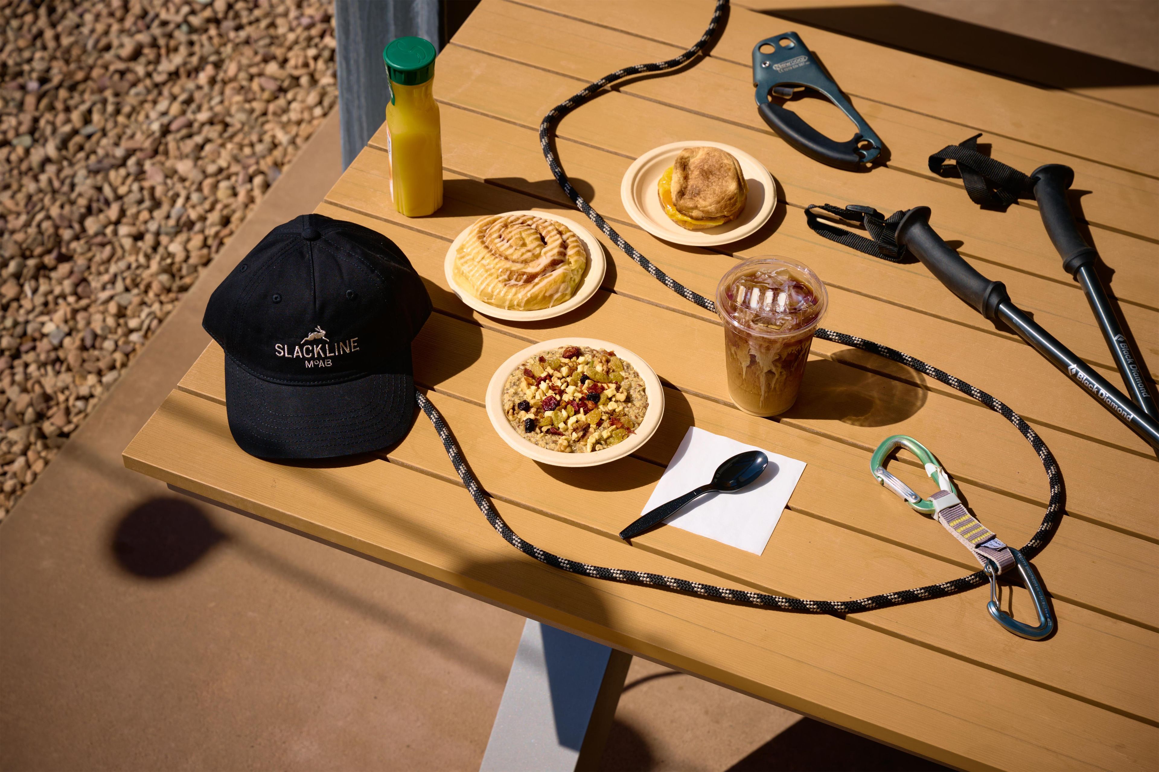 A wooden table with a black cap, two plates of food, a bottle, a spoon, and a carabiner.