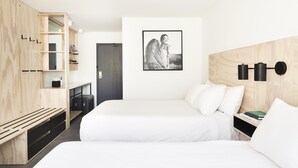 A modern hotel room with two beds, a wooden wardrobe, a black and white artwork on the wall, and a bedside table with a book.