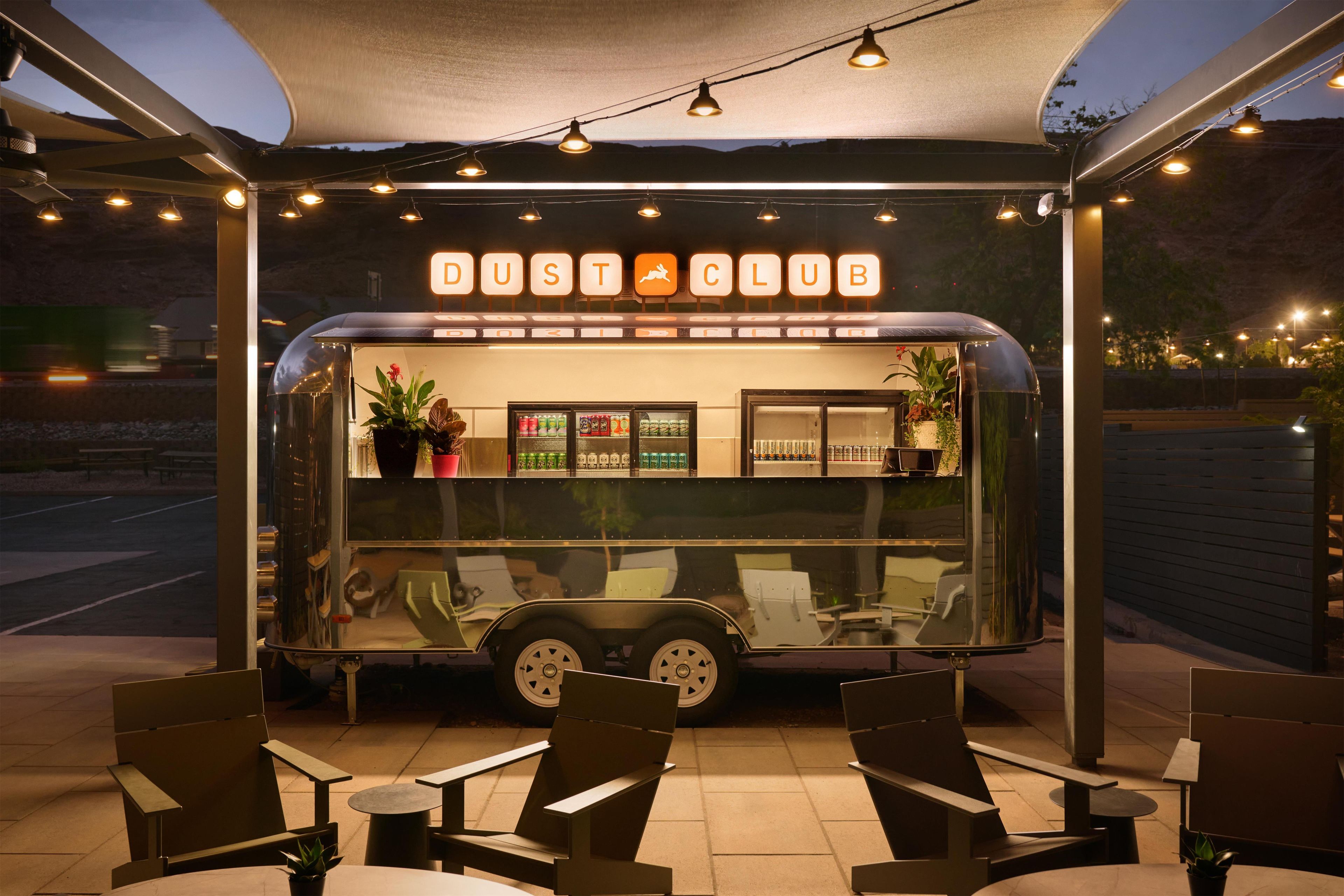Un camion-restaurant avec une enseigne indiquant « DUST CLUB » est garé sous un auvent décoré de guirlandes lumineuses. On y trouve un présentoir avec des boissons et un espace aménagé avec des chaises et des tables.