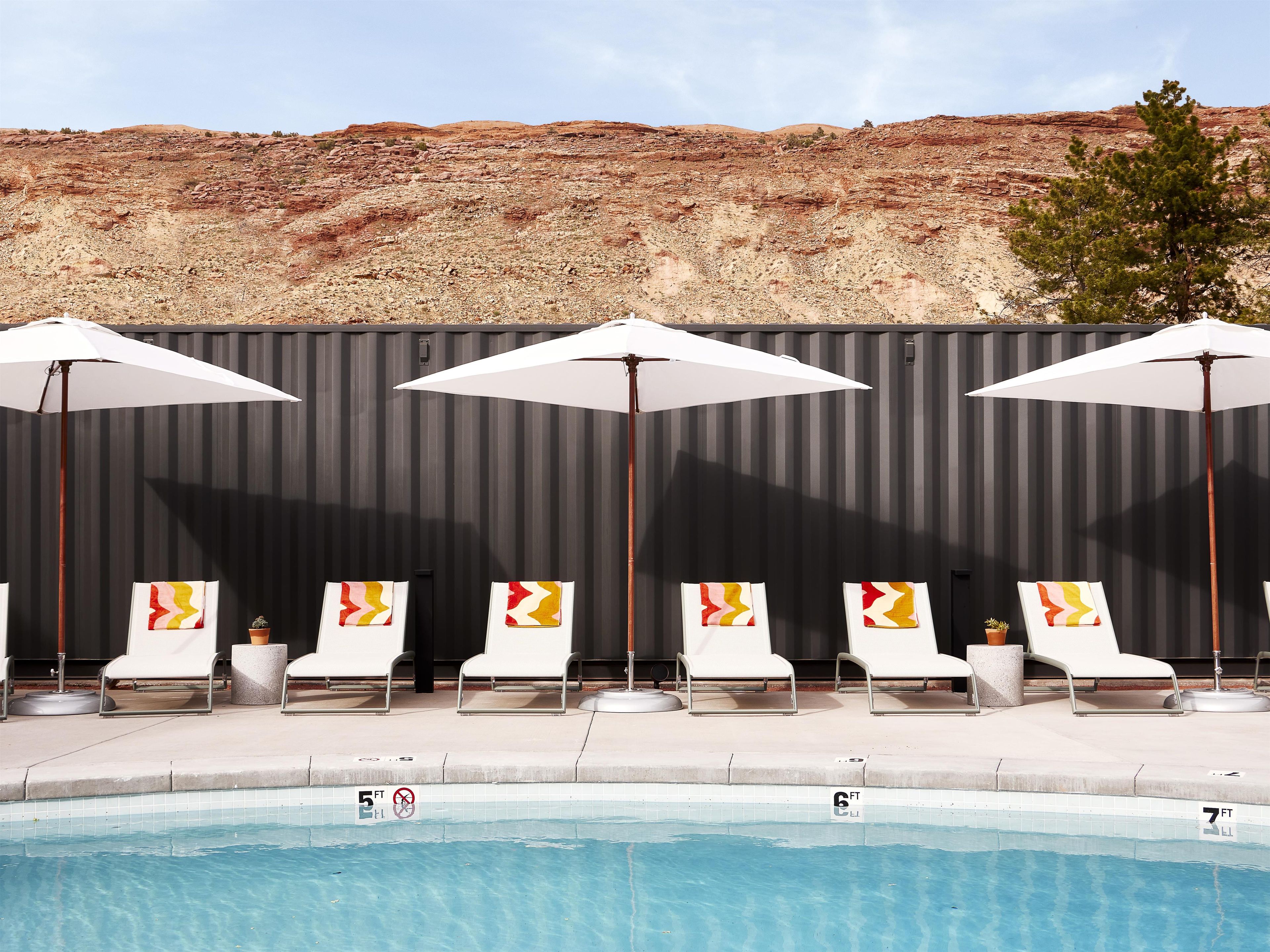 A poolside area with lounge chairs and umbrellas, a tiled pool with depth markers, and a rocky hillside in the background.