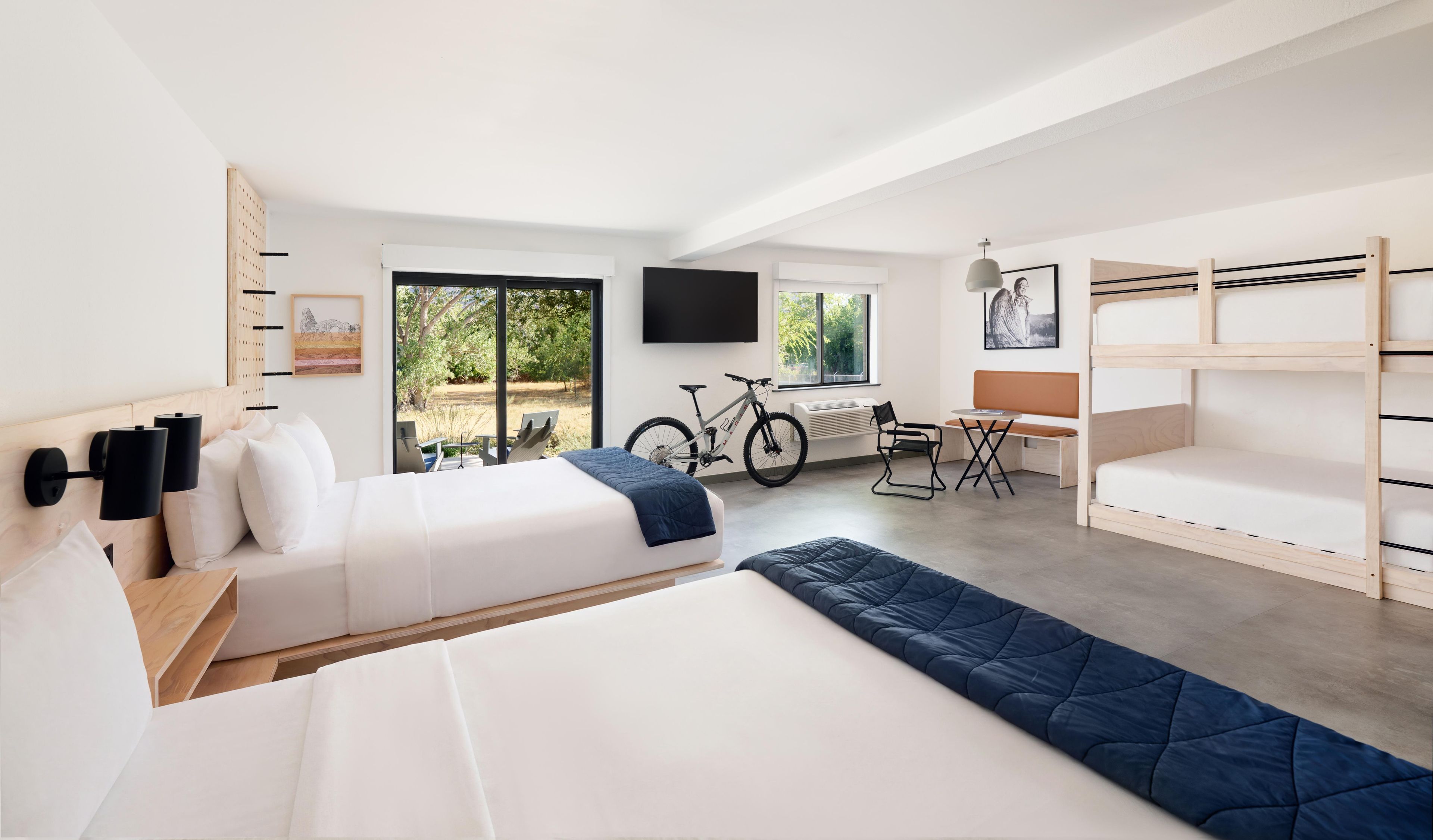 A modern hotel room with a bed, a desk, a bicycle, and a view of the outdoors.