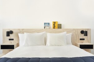 Una cama bien hecha con sábanas blancas y dos almohadas, flanqueada por mesitas de noche de madera con apliques de pared negros, y un cabecero de madera con un diseño sencillo.