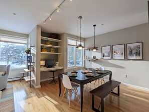 Een moderne eetruimte met een houten tafel, stoelen en een bank. Er zijn planken met decoratieve objecten en een computer op een bureau.