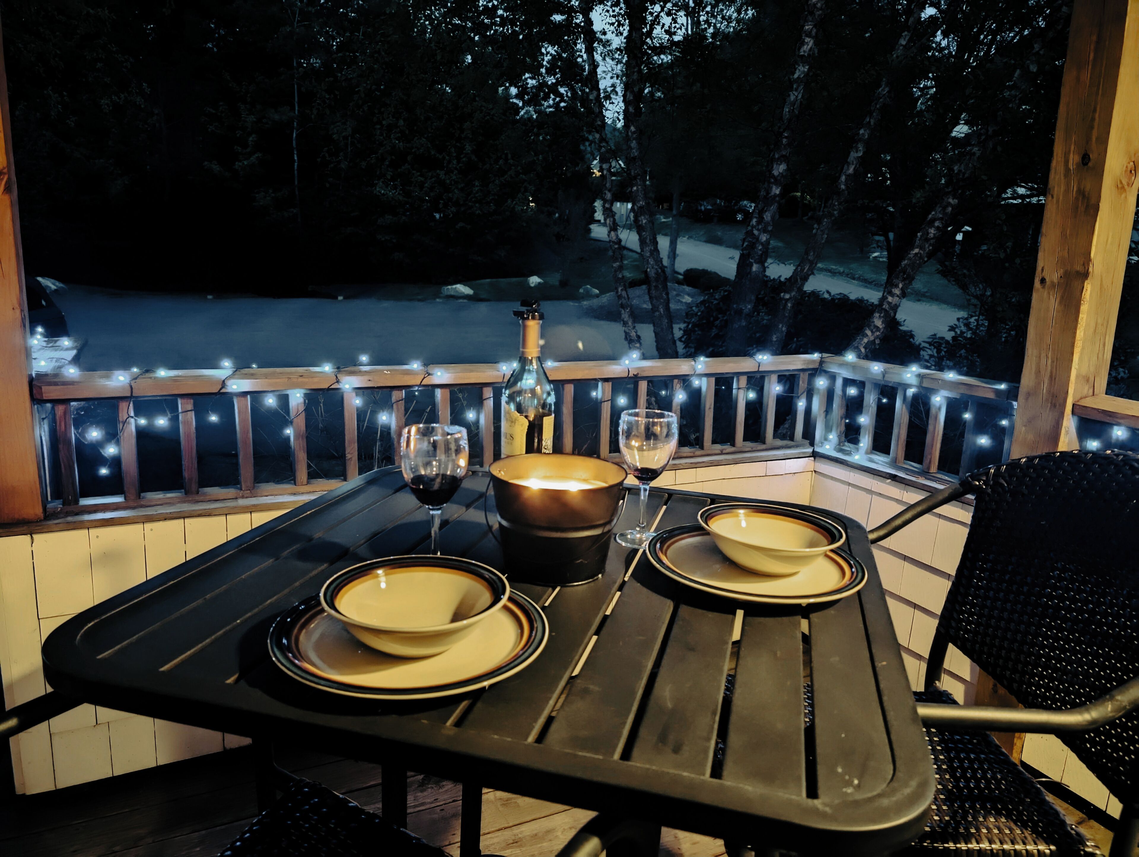Mesa preparada para dos con copas de vino, una botella y cuencos en una terraza con luces colgantes.