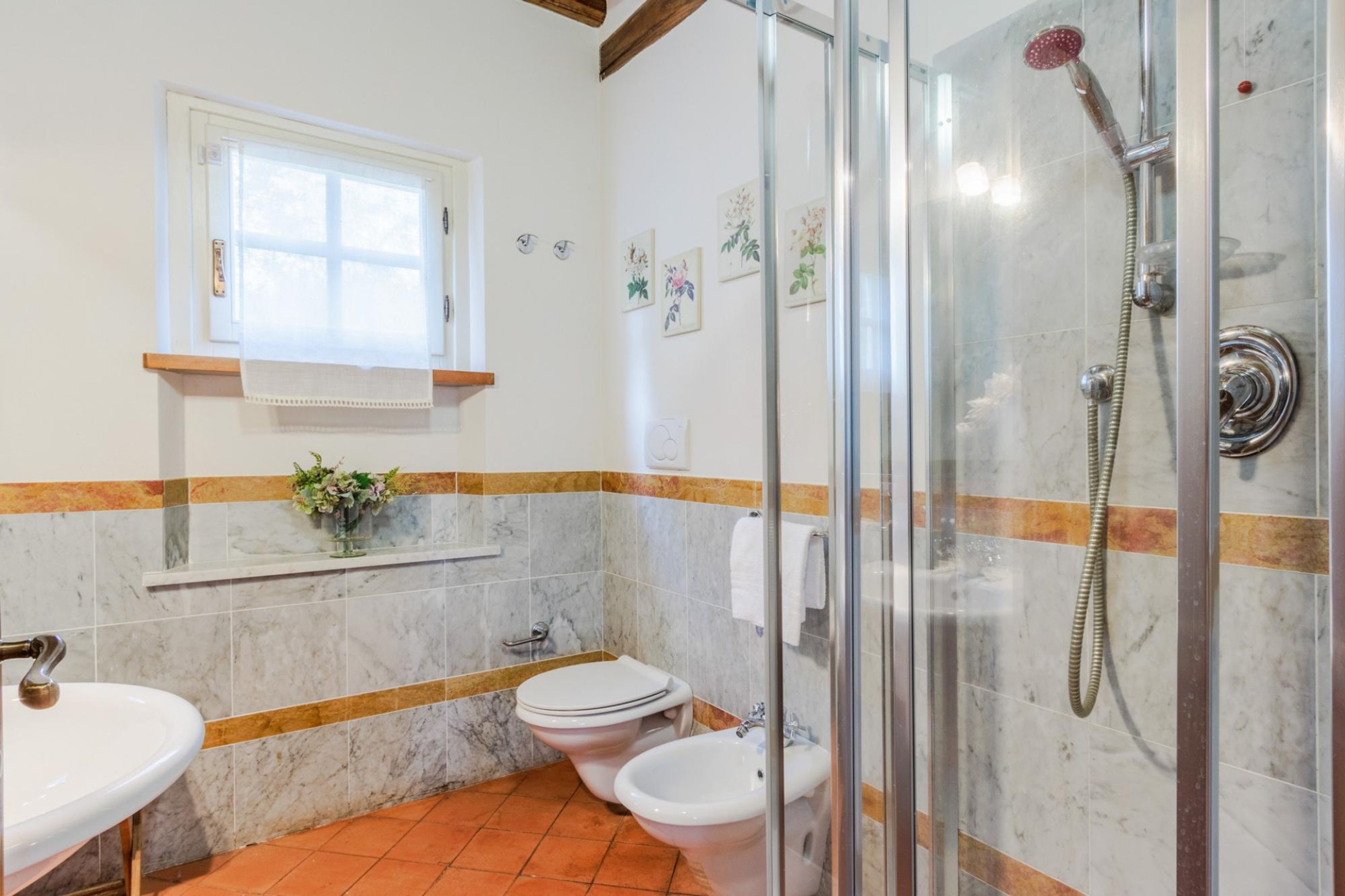 A bathroom with a glass-enclosed shower, two toilets, a sink, and a window with a towel hanging.