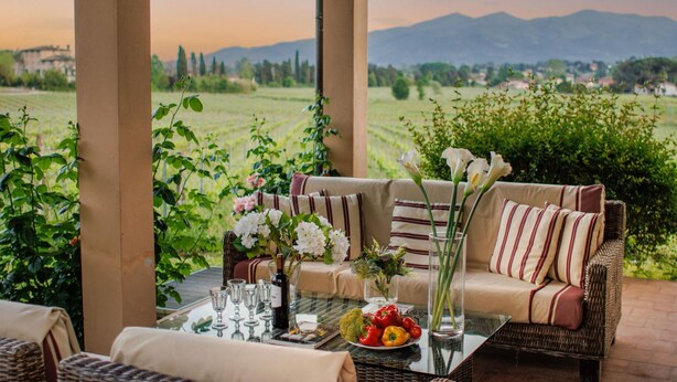 An outdoor seating area with wicker furniture, a glass table, and a view of a vineyard and mountains. - Cicale Farmhouse 3 Bedrooms Farmhouse With Pool (Lucca)