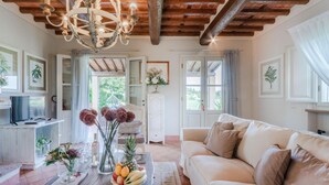 A cozy living room with a wooden ceiling, a chandelier, a sofa, a coffee table with a fruit bowl, and a television. - Cicale Farmhouse 3 Bedrooms Farmhouse With Pool (Lucca)