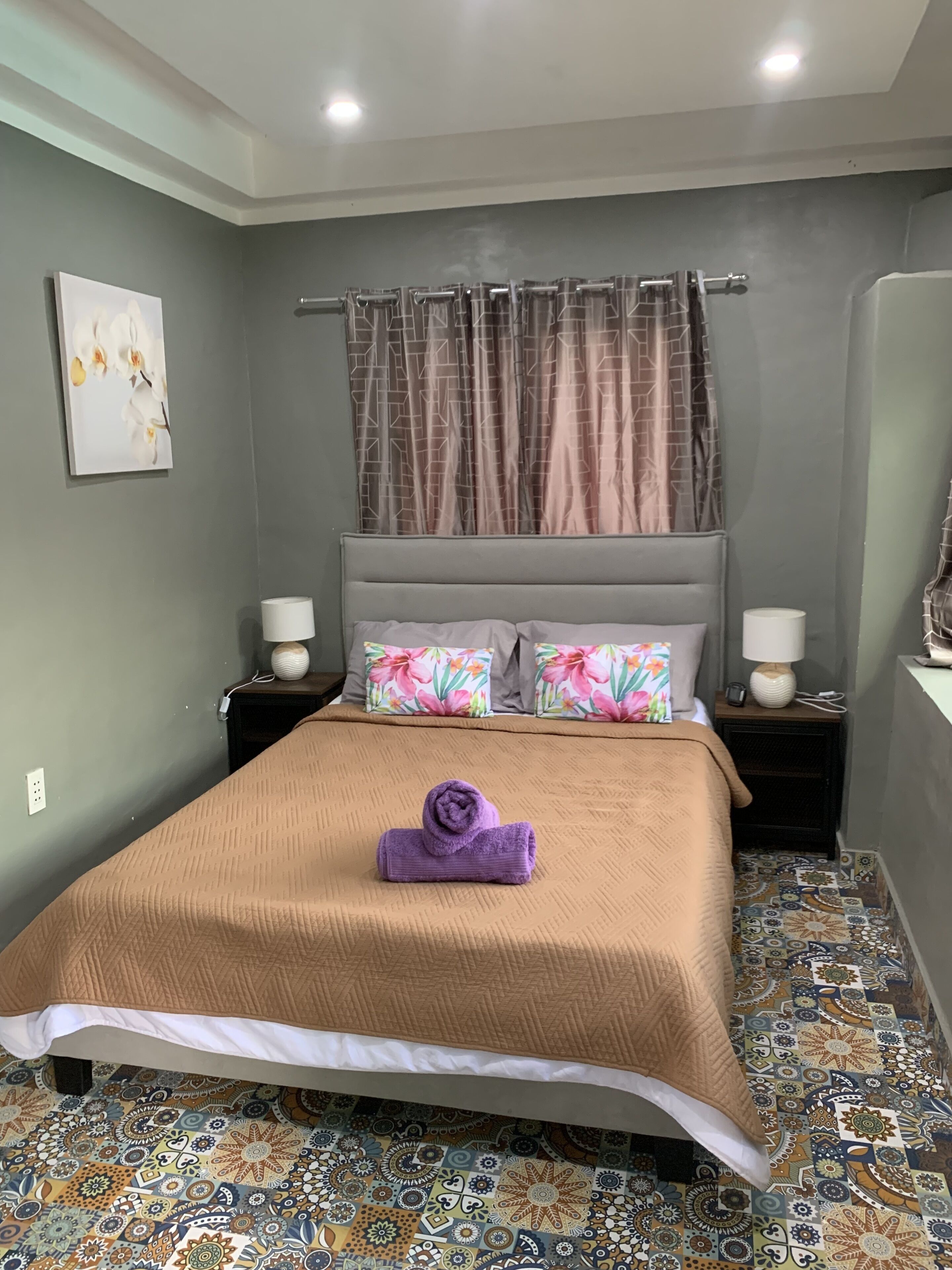 A neatly made bed with a brown bedspread and a purple towel, two floral pillows, and a headboard with a curtain.