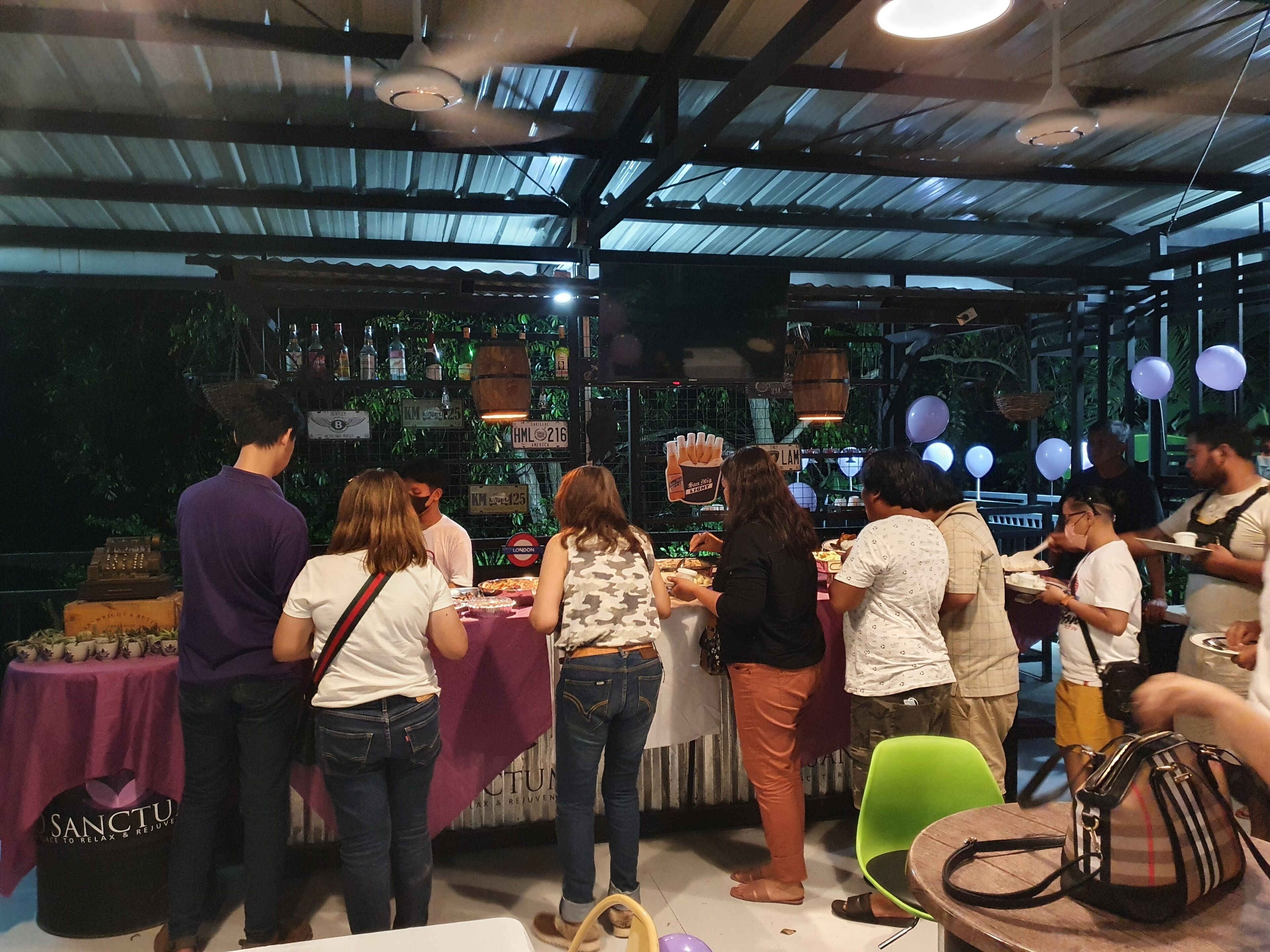 Un grupo de personas cenando en un restaurante al aire libre con techo de metal y mesas cubiertas con manteles morados.