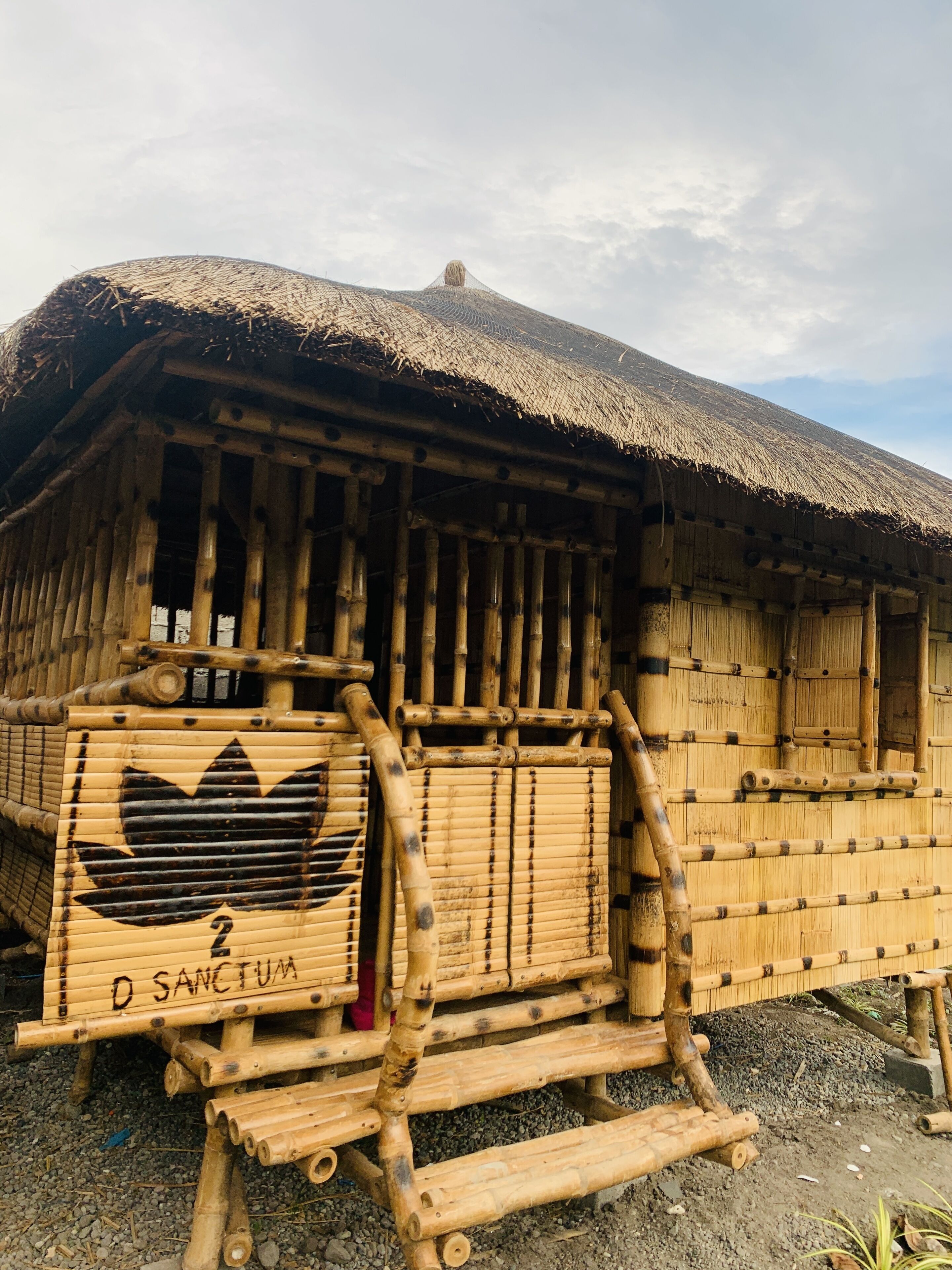 A traditional bamboo hut with a thatched roof, a wooden door, and a sign reading "D SANCTUM".