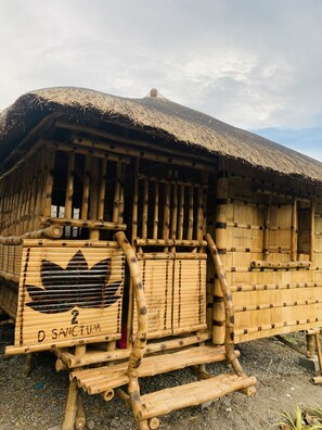 Eine traditionelle Bambushütte mit einem Reetdach, einer Holztür und einem Schild mit der Aufschrift 'D SANCTUM'.