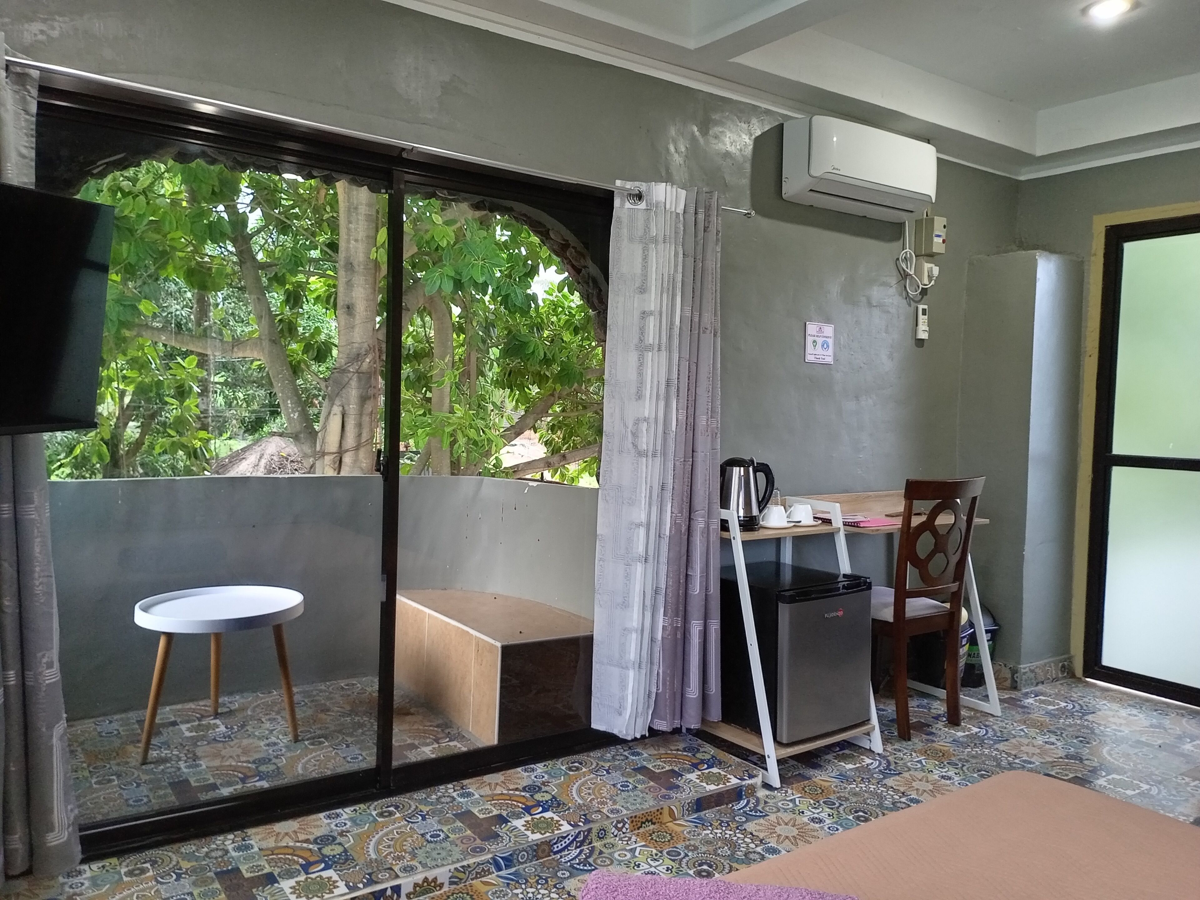 A room with a sliding glass door leading to a balcony, a small table with a chair, a black refrigerator, and an air conditioning unit on the wall.