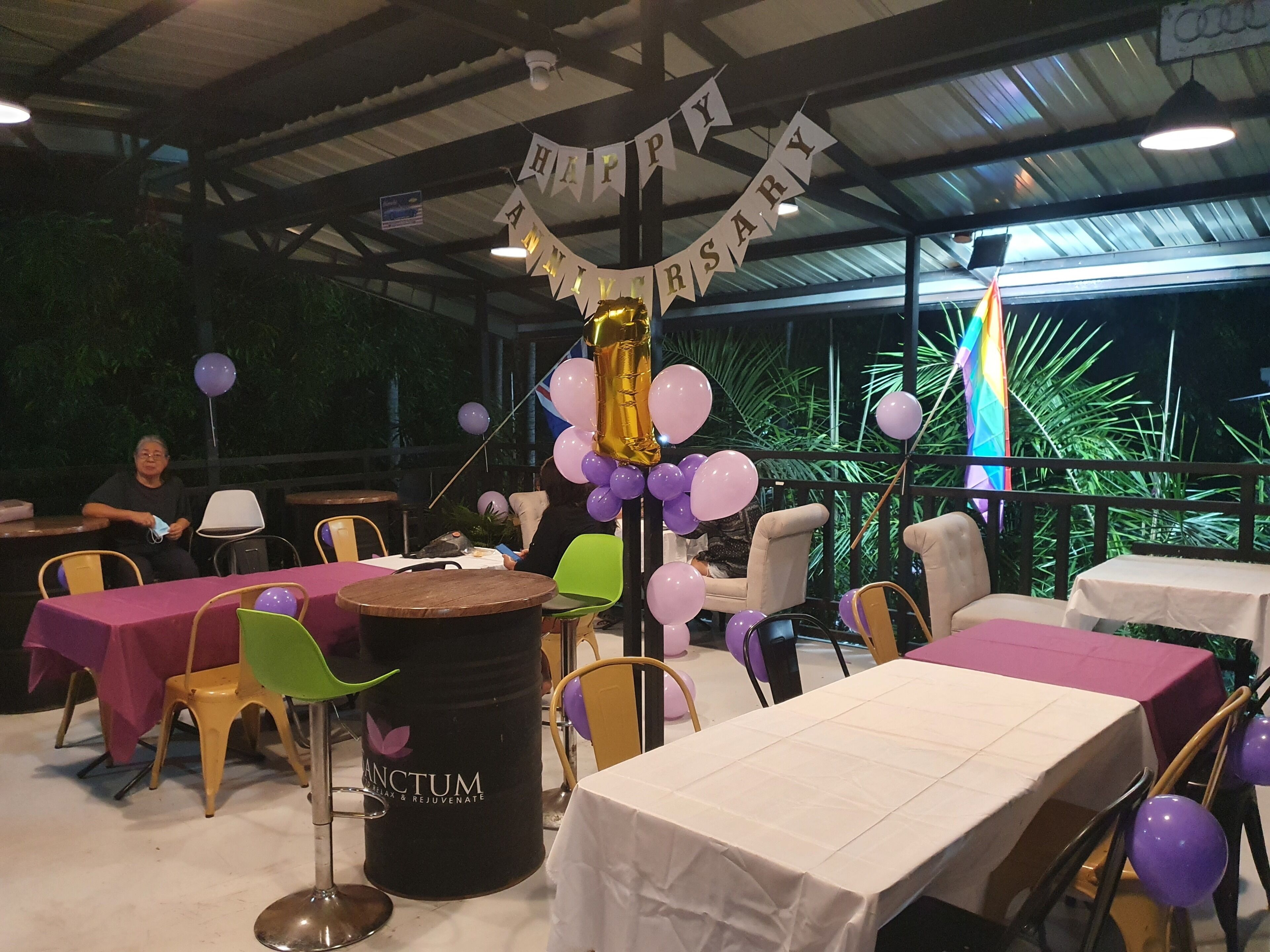 An indoor event space with tables and chairs, a 'Happy Anniversary' banner, balloons, and a barrel with 'ANCTUM' branding.