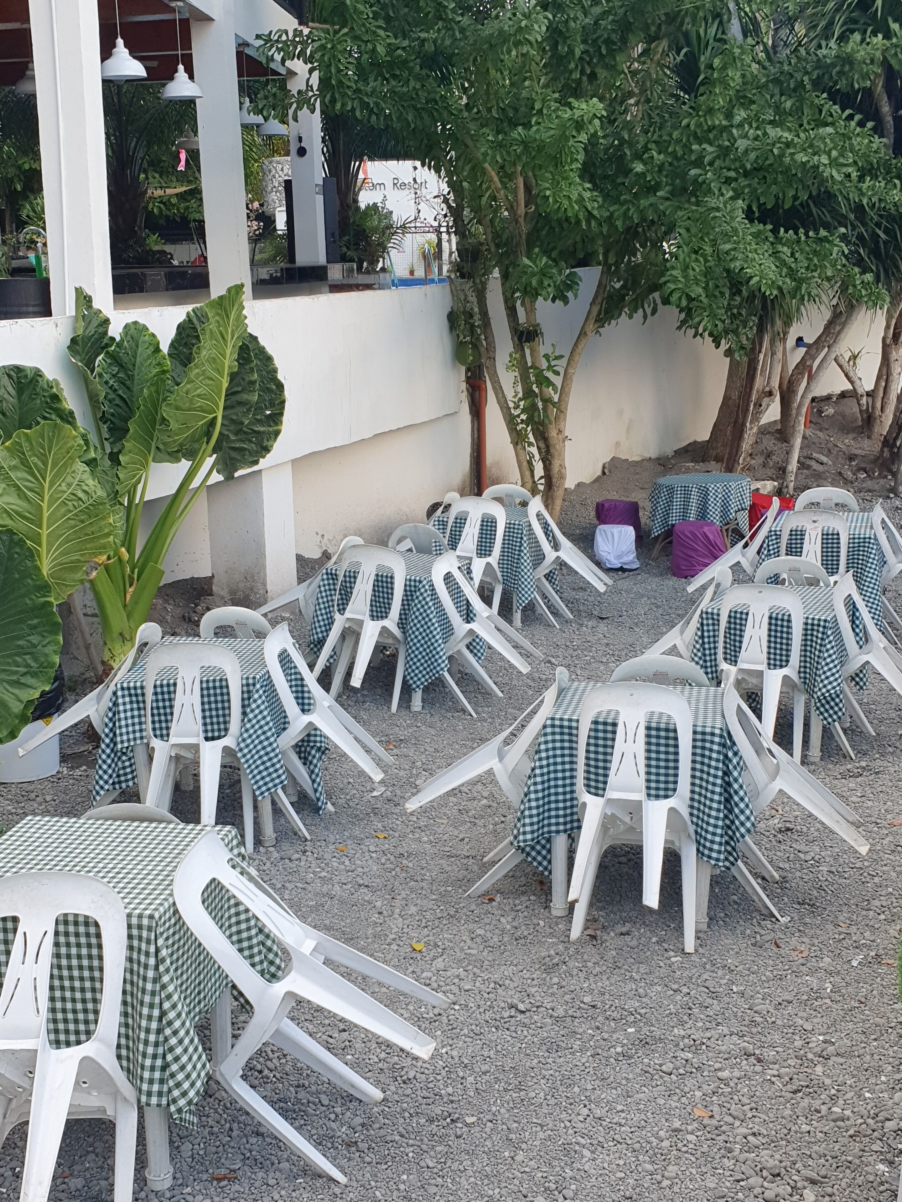 Outdoor seating with white plastic chairs and tables, some covered with green and white checkered tablecloths, surrounded by gravel and greenery.