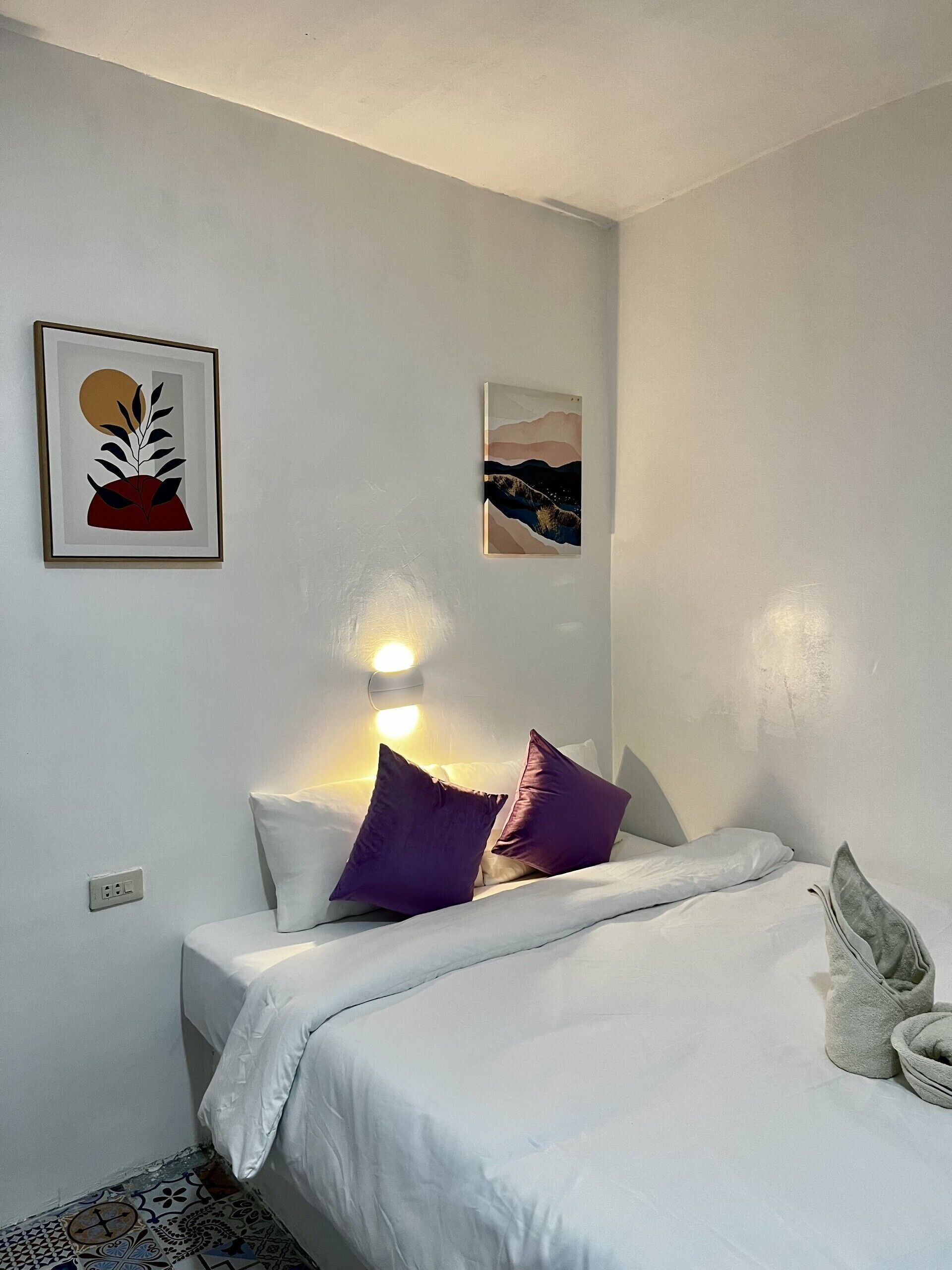 Habitación de hotel con cama, dos almohadas, una lámpara de pared y dos obras de arte enmarcadas.