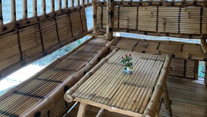 Estructura de bambú con techo de paja, zonas de estar y una mesa con un florero.
