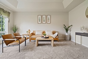 Un salon comprenant un canapé, deux fauteuils, une table basse et une table d’appoint. Trois reproductions botaniques encadrées ornent le mur, ainsi qu’une plante en pot et un miroir.