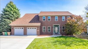 A two-story brick house with a front garage, a well-maintained lawn, and a large tree to the left. - Home w/Hot Tub -Spacious, Quiet Neighborhood Near Des Moines, IA. (Clive)
