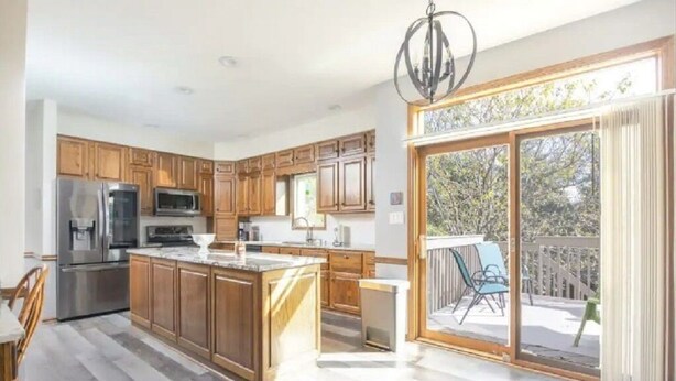 A spacious kitchen with wooden cabinets, a central island, and a sliding glass door leading to an outdoor area with a deck and chairs. - Home w/Hot Tub -Spacious, Quiet Neighborhood Near Des Moines, IA. (Clive)