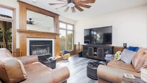 A spacious living room with leather sofas, a fireplace, a large TV, and a ceiling fan. - Home w/Hot Tub -Spacious, Quiet Neighborhood Near Des Moines, IA. (Clive)