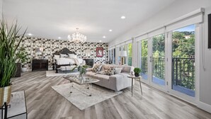 Un salon spacieux avec un grand canapé, une table basse et un lustre. Au fond, on aperçoit un lit avec une tête de lit, et un balcon d’où l’on peut admirer les arbres et les maisons.