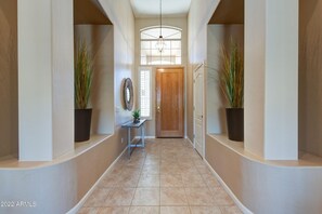 Stroll down this serene hallway, lined with lush greenery and natural light, featuring a wooden door and a stylish light fixture.