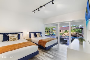 Two large beds with blue headboards and white bedding, a wooden bedside table, and a sliding glass door to an outdoor area with a pool.