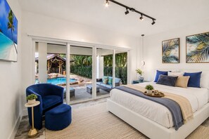 Une chambre dotée d’un grand lit, d’un coin salon avec un fauteuil et une petite table, et offrant une vue sur une piscine extérieure et un jardin.