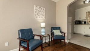 Une pièce comprenant deux fauteuils en cuir bleu, une petite table avec une lampe et un coin cuisine avec des meubles blancs.